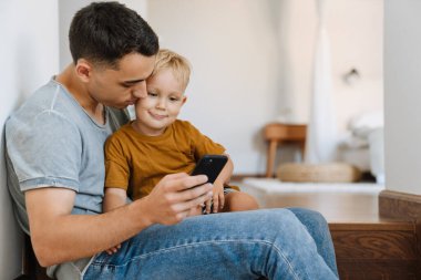 Evde merdivenlerde otururken cep telefonu kullanan genç bir baba ve oğul.
