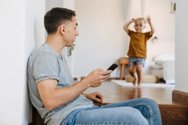 Genç baba oğluyla evde vakit geçirirken cep telefonu kullanıyor.