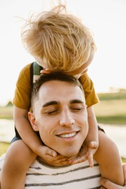 Genç beyaz baba dışarıda yürürken küçük oğlunu omuzlarında taşıyor.