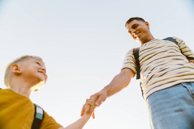 Genç beyaz baba ve küçük oğlu dışarıda yürürken gülümsüyor ve el ele tutuşuyorlar.