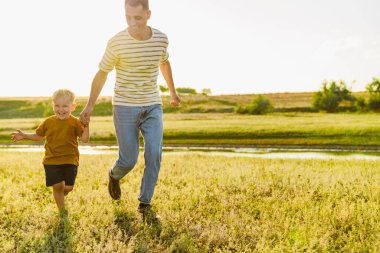 Genç beyaz bir baba ve küçük oğlu yaz tarlasında koşarken el ele tutuşuyorlar.