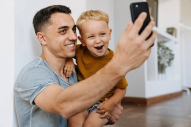 Evde cep telefonuyla gülümseyen ve selfie çeken genç baba ve oğul.