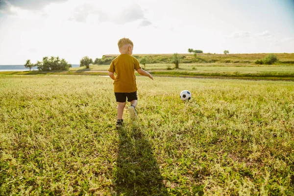 Sarışın küçük beyaz çocuk sahada koşuyor ve dışarıda futbol oynuyor.
