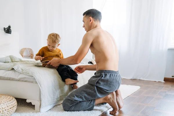 Young Shirtless Father Reading Book While Dressing His Son Home — стоковое фото