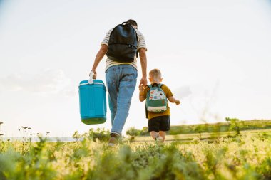 Genç beyaz baba ve oğul yaz tarlasında çantalarla yürüyorlar.