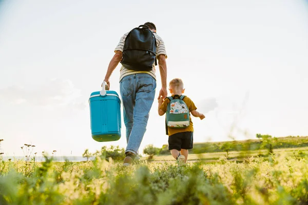 Genç beyaz baba ve oğul yaz tarlasında çantalarla yürüyorlar.
