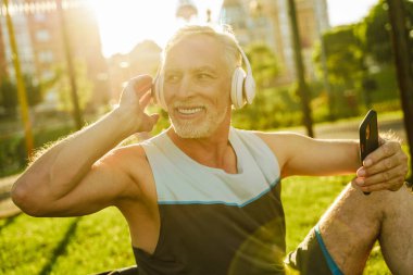 Gri adam dışarıda çimenlerde otururken müzik dinliyor ve cep telefonu kullanıyor.