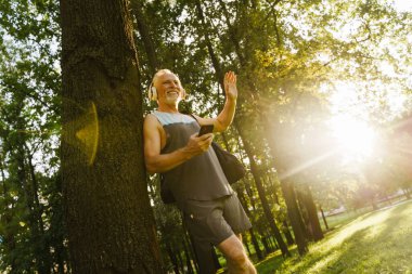 Park yerinde cep telefonu kullanırken gülümseyen ve el sallayan gri kıdemli adam.