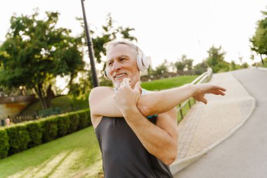 Gri kıdemli adam parkta çalışırken müzik dinliyor ve egzersiz yapıyor.