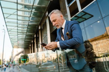 Takım elbise giymiş üst düzey bir adam şehir caddesinde beklerken cep telefonu kullanıyor.