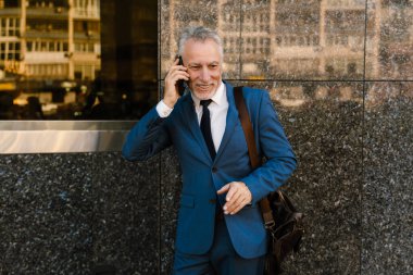 Takım elbise giymiş üst düzey bir adam şehir caddesinde beklerken cep telefonuyla konuşuyor.