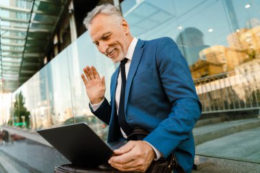 Belediye binasında otururken el sallayan ve dizüstü bilgisayar kullanan kıdemli bir adam.