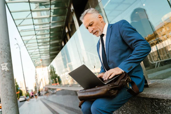 Takım elbise giymiş üst düzey bir adam şehir caddesinde otururken dizüstü bilgisayarla çalışıyor.