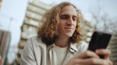 Mutlu kıvırcık saçlı adam bankta otururken cep telefonuna bakıyor.