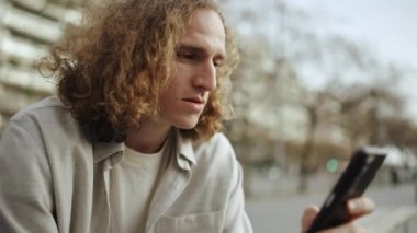 Güzel kıvırcık saçlı adam bankta otururken cep telefonuna bakıyor.