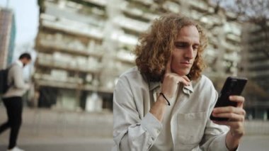 Dalgın kıvırcık saçlı adam bankta otururken cep telefonuna bakıyor.