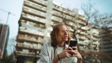 Meditasyon yapan kıvırcık saçlı adam bankta otururken cep telefonuna bakıyor.