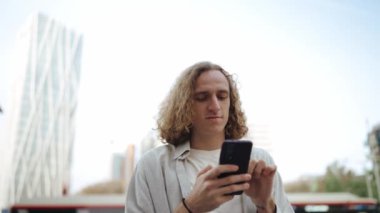 Dalgın kıvırcık saçlı adam dışarıda telefonla yazı yazıyor.