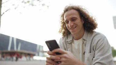Cheerful curly-haired man texting by mobile while sitting on the bench outdoors