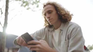 Yakışıklı kıvırcık saçlı adam dışarıda otururken telefonla yazı yazıyor.