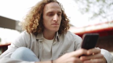 Dalgın kıvırcık saçlı adam dışarıda otururken telefonla yazı yazıyor.