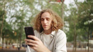 Açık havada otururken telefonla konuşan kıvırcık saçlı olumlu bir adam.