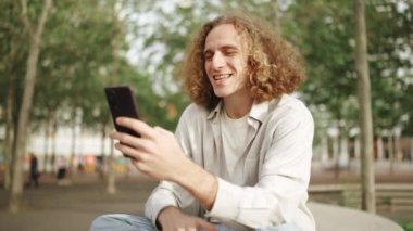 Gülümseyen kıvırcık saçlı adam dışarıda otururken telefonla konuşuyor.