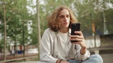 Mutlu kıvırcık saçlı adam dışarıda otururken telefonla konuşuyor.