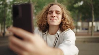 Dışarıda otururken telefonla konuşan kıvırcık saçlı, gülen adam.