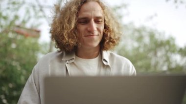 Gülümseyen kıvırcık saçlı adam dışarıda otururken dizüstü bilgisayarda çalışıyor.