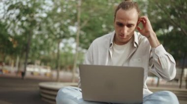 Ciddi kıvırcık saçlı adam dışarıda otururken dizüstü bilgisayarla çalışıyor.