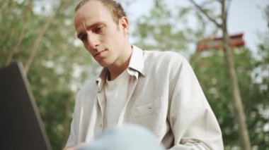 Kendine güvenen kıvırcık saçlı adam dışarıda otururken dizüstü bilgisayarda çalışıyor.