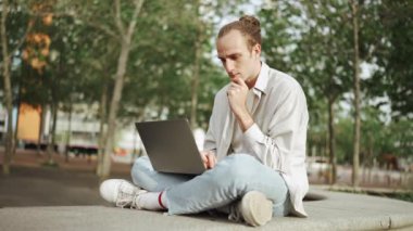 Yoğunlaşmış kıvırcık saçlı adam dışarıda otururken dizüstü bilgisayarda çalışıyor.