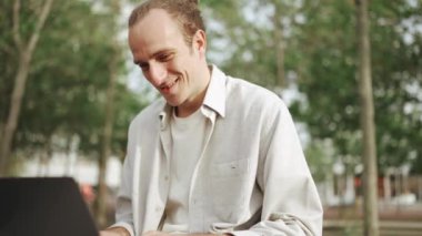Gülen kıvırcık saçlı adam dışarıda otururken dizüstü bilgisayarda çalışıyor.