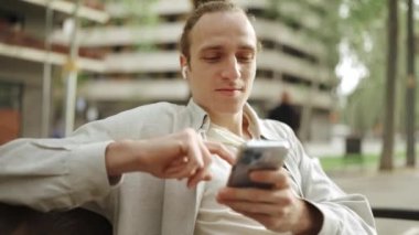 Kulaklıkla müzik dinleyen ve dışarıda telefonla mesajlaşan kıvırcık saçlı olumlu bir adam.