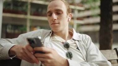 Neşeli kıvırcık saçlı adam kulaklıkla müzik dinliyor ve dışarıda telefonla mesajlaşıyor.