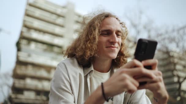 Smiling Curly Haired Man Typing Mobile While Sitting Bench Outdoors — Video Stock