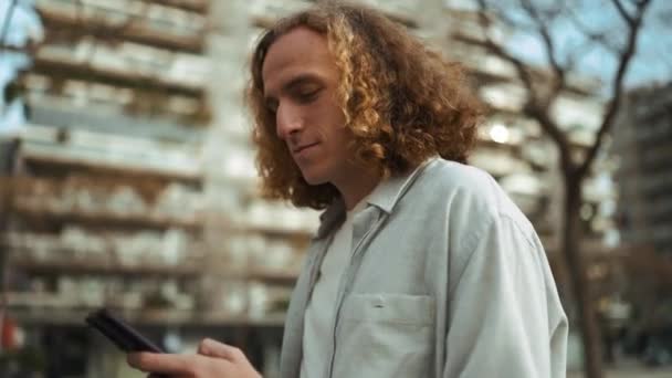 Pretty Curly Haired Man Looking Mobile While Walking Outdoors — Vídeo de Stock