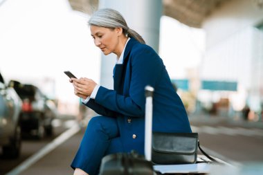 Havaalanının yanındaki bankta otururken cep telefonu kullanan gri Asyalı kadın.