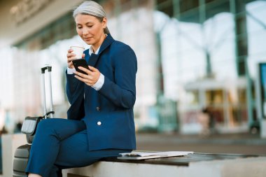 Gri Asyalı kadın hava alanında cep telefonu kullanıyor ve kahve içiyor.