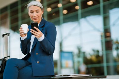 Gri Asyalı kadın hava alanında cep telefonu kullanıyor ve kahve içiyor.