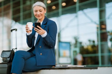 Gri Asyalı kadın hava alanında cep telefonu kullanıyor ve kahve içiyor.