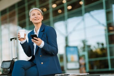 Gri Asyalı kadın hava alanında cep telefonu kullanıyor ve kahve içiyor.