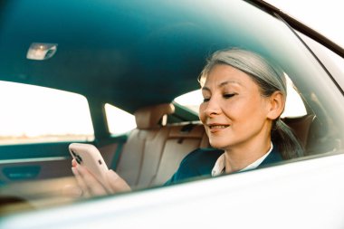 Olgun Asyalı iş kadını araba kullanırken cep telefonu kullanıyor.