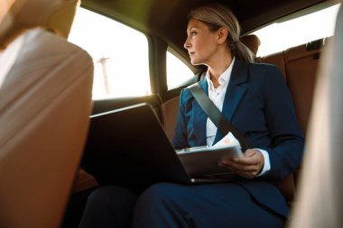 Olgun Asyalı iş kadını arabada otururken laptop ve kağıtlarla çalışıyor.