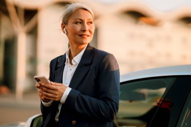 Olgun Asyalı kadın arabada beklerken kulaklık ve cep telefonu kullanıyor.