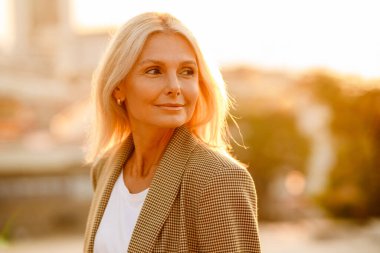 Olgun beyaz iş kadını takım elbiseli, gülümseyen ve dışarı bakan.