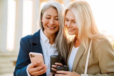 Olgun, çok ırklı iş kadınları dışarıda buluşurken cep telefonu kullanıyor ve kahve içiyor.