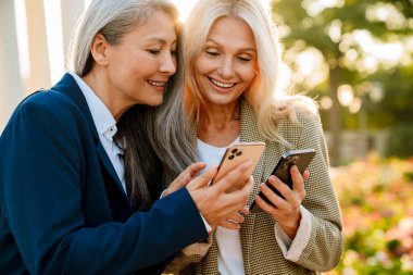 Açık havada buluşurken gülümseyen ve cep telefonu kullanan çok kültürlü iş kadınları.