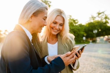Açık havada buluşurken gülümseyen ve cep telefonu kullanan çok kültürlü iş kadınları.